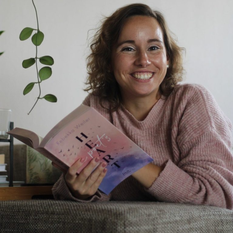 Paket Heartbeat Frau mit lockigem Haar hält ein Buch und lächelt, im Hintergrund Pflanzen sichtbar.
