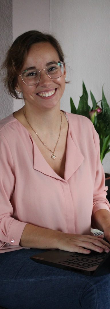 Lektorin bei der Arbeit Lächelnde Frau mit Brille, die am Tisch an einem Laptop arbeitet, umgeben von Pflanzen.