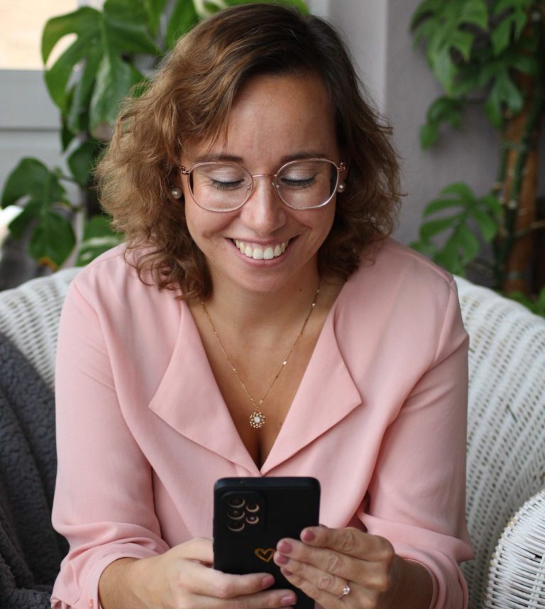 Welches Angebot passt zu mir? Frau mit Brille in rosa Bluse, lächelnd und auf ein Smartphone schauend.